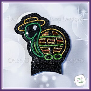 May include: A black glitter turtle with a green and yellow shell, wearing a yellow hat. The turtle is holding a green and yellow globe.