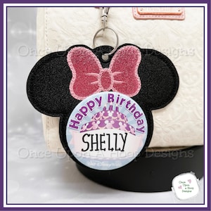 May include: A black felt Minnie Mouse head with a pink glitter bow and a round blue button with the text "Happy Birthday SHELLY" and a purple and pink cake with candles. The button also has the text "Walt Disney World" at the bottom.