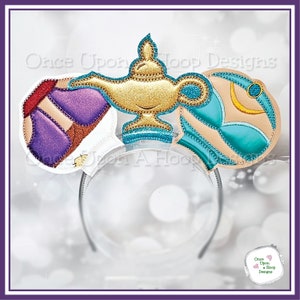 May include: A decorative headband featuring Aladdin-themed designs. The headband has a clear plastic band with attached fabric cutouts. The cutouts include a gold genie lamp, purple and tan shapes, and teal and tan shapes. The design is embellished with glitter.