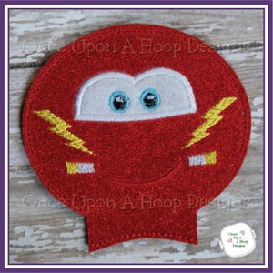 May include: Red felt applique of a cartoon car with a white face, blue eyes, and yellow lightning bolts. The applique is round and has a glittery texture.