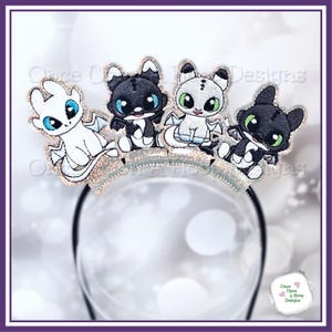 May include: A black headband featuring four embroidered dragon characters in white, black, and gray with glitter accents. The dragons have blue and green eyes and are arranged across a silver glittery band. The text "Once Upon a Hoop Designs" is visible.