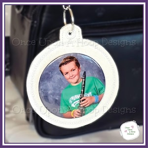 Puede incluir: Un llavero fotográfico circular blanco con una foto de una persona sosteniendo un clarinete. El llavero tiene un borde decorativo cosido y un anillo de metal plateado para sujetarlo. La persona de la foto lleva una camiseta verde.