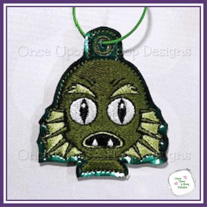 Puede incluir: Un colgante bordado verde con un diseño de criatura, ojos blancos y boca ancha. Tiene un cordón verde para colgar. El texto "Once Upon a Hoop Designs" es visible.