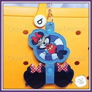Puede incluir: Un personaje de Minnie Mouse azul y blanco con gafas de sol está bordado en un círculo azul y blanco. El círculo está unido a un amuleto azul y blanco con un par de gafas de sol negras con lazos de lunares rojos y blancos.