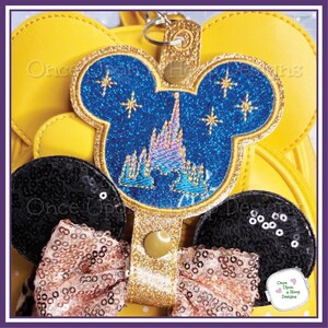 May include: A Mickey Mouse-shaped keyring with a blue glitter design featuring a castle and stars. The keyring has a gold border and strap. The image also shows black sequined ears and a pink sequined bow.