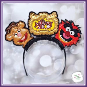 Puede incluir: Una diadema negra con personajes de The Muppet Show. La diadema tiene un letrero con marco dorado con el logotipo del programa y recortes de personajes de Fozzie Bear y Animal. Los personajes son en tonos marrón, rojo y negro.