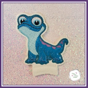 May include: Embroidered patch of a blue lizard with purple accents and large blue eyes. The lizard is standing on a white background. "Once Upon A Hoop Designs" is written on a white circle in the bottom right corner.