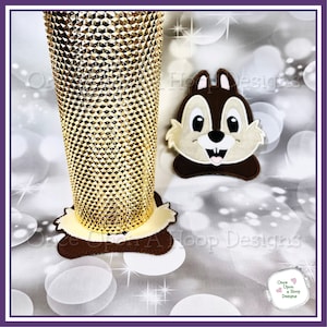 May include: A brown and beige embroidered chipmunk face applique with a white background. The applique is sitting on a white surface with a gold glitter tumbler behind it.