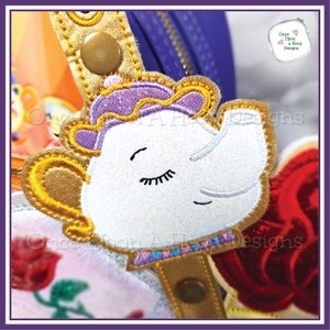 May include: A white teapot with a gold rim and handle, purple lid, and closed eyes. The teapot is embroidered with glitter and is attached to a gold strap.