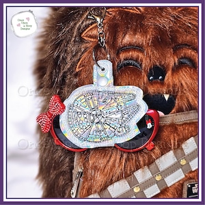 Op de afbeelding: Een zilveren en iriserende sleutelhanger met een Millennium Falcon-ontwerp. De sleutelhanger is bevestigd aan een paar rode zonnebrillen met een rode strik en een Mickey Mouse-kopbedel.