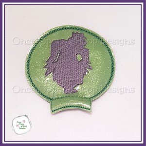May include: A green and purple embroidered patch featuring a silhouette of a figure inside a snow globe. The patch has a glittery green background and a stitched border. The text "Once Upon a Hoop Designs" is visible.