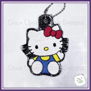 May include: A white embroidered Hello Kitty keychain with a red bow, blue overalls, and yellow trim. The keychain has a black border and a silver chain. The text "Once Upon a Hoop Designs" is visible.