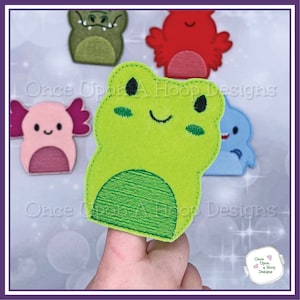 May include: A collection of felt finger puppets in various animal designs. The primary focus is on a green frog puppet with embroidered details. Other puppets include a red creature, a pink axolotl, a blue dolphin, and a green alligator. The text "Once Upon A Hoop Designs" is visible.