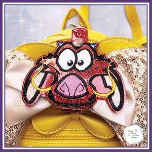 May include: A red and black embroidered keychain featuring a cartoon warthog character with large eyes and a pink snout. The keychain is attached to a yellow backpack with a bow and sequined accents. The text "Once Upon a Hoop Designs" is visible.