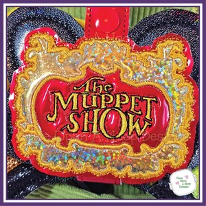 May include: A red, gold, and iridescent patch featuring the text "The Muppet Show." The patch has a gold embroidered border and a red background. The patch is attached to a black and green background.