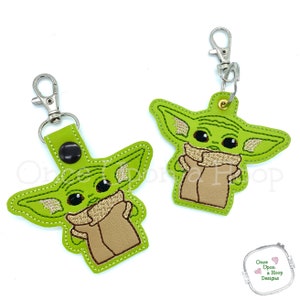 May include: Two green embroidered keychains featuring a cartoon character with large eyes and ears. The character is holding a brown object in its arms. The keychains are attached to silver key rings.