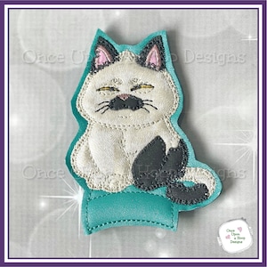 May include: A white and black cat finger puppet with a teal base. The cat is sitting with its paws crossed and has a grumpy expression.