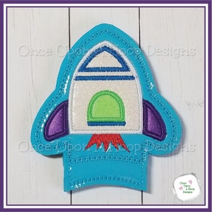 Può includere: Un'applicazione a razzo blu e bianca con accenti viola e rossi. Il razzo è ricamato con glitter e ha un contorno bianco. L'applicazione è perfetta per aggiungere un tocco divertente e fantasioso a qualsiasi progetto.