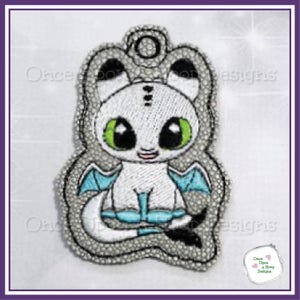 May include: Embroidered white dragon with large green eyes, small blue wings, and a black tail. The dragon is outlined in gray and has a small loop at the top for hanging. The design is on a white background with a purple border.