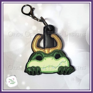 May include: A green and gold alligator head keychain with a black loop for attaching to a bag or keys. The alligator has a toothy grin and gold horns.