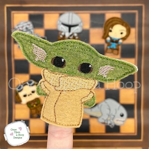 May include: Green and brown felt finger puppet of a character from the Star Wars franchise, "The Mandalorian". The puppet is on a finger with a checkerboard background.