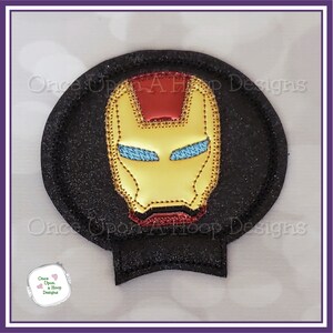 May include: A black and gold iron man mask embroidered on a black circle with glitter. The mask has a red forehead piece and blue eyes.