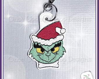 May include: A green Grinch keyring with a red Santa hat and white trim. The keyring has a silver clasp and a white tag. The Grinch's face features yellow eyes and a mischievous grin. The text "Once Upon a Hoop Designs" is visible.