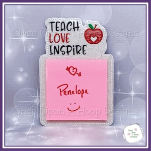 Könnte beinhalten: Ein weißer Glitzer-Notizblockhalter mit einem roten Apfel und dem Text "TEACH LOVE INSPIRE". Der Notizblock ist rosa und hat den Namen "Penelope" darauf geschrieben.