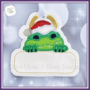 May include: A green alligator wearing a Santa hat and peeking over a white sign with the text "Once Upon A Hoop Designs".