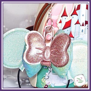 以下が含まれることがあります： ピンク色のキラキラ光るリボン型のキーホルダー。中央は薄いピンク色で、シルバーの留め金が付いています。リボンは水色と虹色の生地に付けられています。背景にはお城のイラストが描かれています。