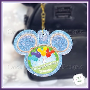 May include: A light blue glittery keychain in the shape of Mickey Mouse ears. The center circle features colorful balloons and the text "I'm Celebrating! Disneyland". The keychain is attached to a gold chain.