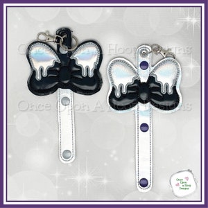 May include: Two black and silver iridescent bow keychains with a white strap. The bows have a dripping effect. The keychains are attached to the straps with silver rings.