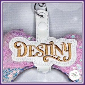 May include: A white leather keychain with the name "Destiny" in gold script. The keychain is attached to a sparkly purple bow with sequins. The design includes the text "Once Upon a Hoop Designs".