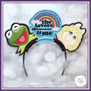 May include: A black headband featuring Kermit the Frog and Miss Piggy character designs. A blue rainbow design with the text "the lovers the dreamers & me" is centered on the headband. The design is outlined in black glitter.