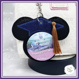 Puede incluir: Un llavero de fieltro azul y negro con forma de oreja de Mickey Mouse con un gorro de graduación y una borla. El llavero presenta una imagen redonda del carruaje de Cenicienta y el castillo con el texto "...Happily Ever After" y "A Hoop World Disney World".