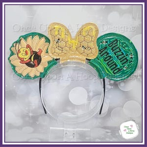 Puede incluir: Orejas de ratón bordadas de color verde y amarillo con un diseño de abeja y girasol. Las orejas tienen el texto "Buzzin' Around" en una oreja y un diseño de panal en la otra.