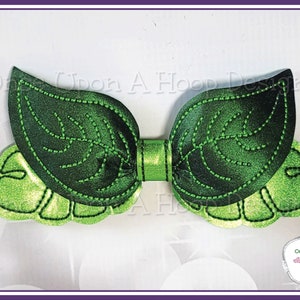 May include: Green fabric bow with a leaf design. The bow is made of two large leaves with a smaller leaf on each side. The leaves have a dark green centre and a lighter green border.