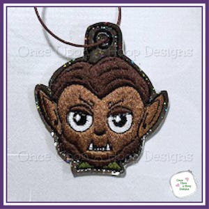 May include: A brown and tan werewolf-themed ornament with a cartoonish design. The ornament features a detailed face with large eyes, pointed ears, and fangs. The text "Once Upon a Hoop Designs" is visible.
