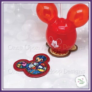 May include: Red Mickey Mouse shaped balloon with a straw and a Mickey Mouse face printed on the front. The balloon is sitting on a brown Mickey Mouse shaped base with a gold design. There is a separate red Mickey Mouse shaped applique with a colorful image of Mickey Mouse, Donald Duck, Goofy, and Pluto.
