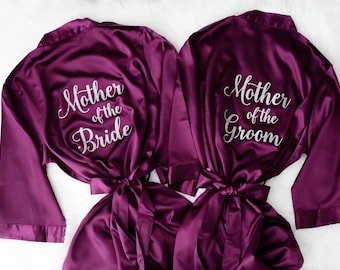 May include: Two deep plum-coloured satin robes with long sleeves and matching waist ties. The left robe features white script text reading Mother of the Bride, while the right robe features white script text reading Mother of the Groom.