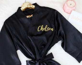 May include: A black satin robe featuring the name Chelsea in gold script lettering on the left chest. The robe has wide sleeves, a matching tie belt at the waist, and is displayed on a gold glitter hanger next to a pink candle and a card.