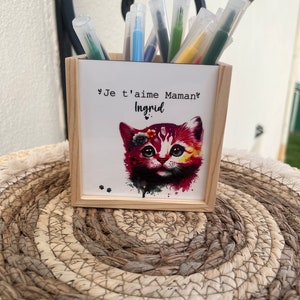 Peut inclure: Un pot à crayons en bois avec une illustration à l'aquarelle d'un chat rouge et jaune. Le pot porte l'inscription "Je t'aime Maman? Ingrid". Il contient divers stylos de couleur et repose sur un tapis tressé.