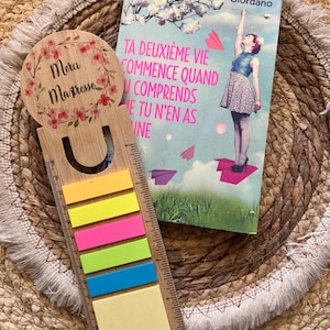 Peut inclure: Un marque-page en bois avec un motif floral et le texte "Mera Maîtresse" à côté d'un livre intitulé "Ta Deuxième Vie Commence Quand Tu N'en As Une". Le marque-page comporte des post-it colorés.