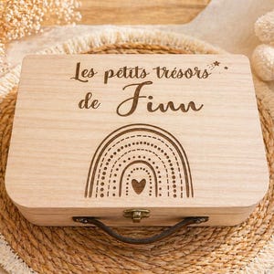 Peut inclure: Un coffret en bois clair avec l'inscription "Les petits trésors de Finn" et un motif arc-en-ciel. La boîte a une poignée et un fermoir. La boîte est posée sur un tapis tissé.