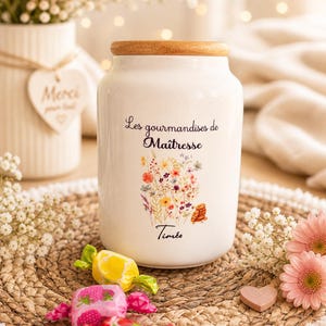 Peut inclure: Un pot en céramique blanc avec un couvercle en bois, orné d'un motif floral et du texte "Les gourmandises de Maîtresse". L'image comprend des bonbons, des fleurs et une décoration en forme de cœur, créant un thème charmant et décoratif.
