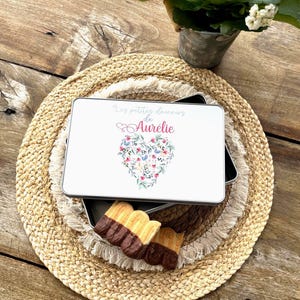 Peut inclure: Une boîte rectangulaire blanche avec le texte "Les petites douceurs de Aurélie" et un motif de cœur floral. La boîte est posée sur un dessous de plat tressé avec des biscuits trempés dans le chocolat. Une petite plante en pot est en arrière-plan.