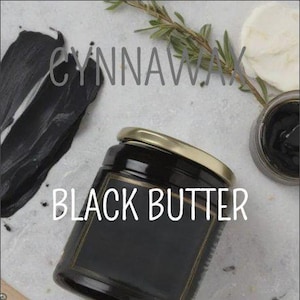 May include: A glass jar with a gold lid filled with black butter. The jar is labelled "Black Butter" and is sitting on a white surface. The text "Soy & Beeswax Melts" is visible in the bottom right corner of the image.
