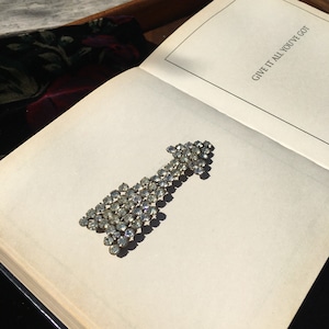 May include: A vintage rhinestone arrow brooch, sparkling with clear crystals, rests on an open book. The brooch is arranged to point upwards. The book's page has the text "GIVE IT ALL YOU'VE GOT".