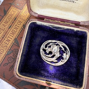 May include: A circular silver brooch featuring an openwork floral design with a central flower motif. The brooch is displayed inside a vintage hinged jewelry box lined with deep purple velvet, featuring the text Edward Anderson Silversmith.