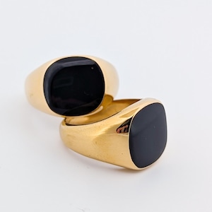 Op de afbeelding: Twee goudkleurige zegelringen met een vierkante, zwarte onyx steen. De ringen worden gepresenteerd op een witte achtergrond, waardoor hun gepolijste oppervlakken worden benadrukt. Klassieke sieraden.